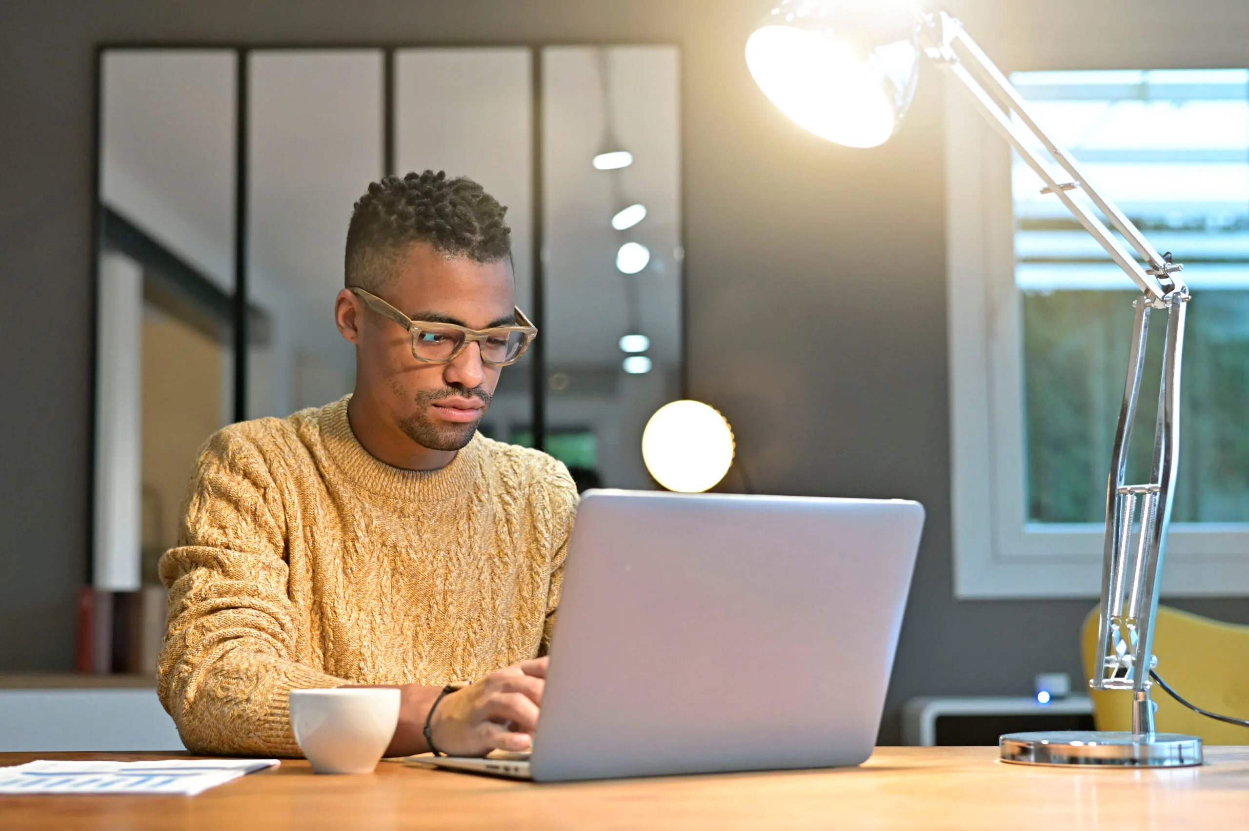 A young man in glasses and a yellow sweater working on a laptop at a desk